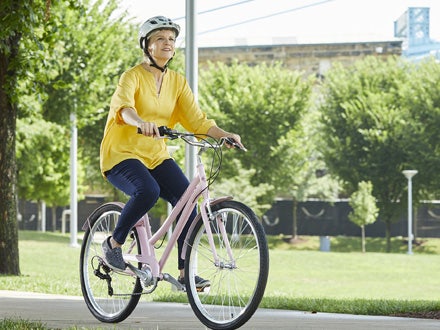 A woman wearing a yellow shirt riding a pink Huffy Sienna bike on a paved path in a park surrounded by trees and greenery. A woman wearing a yellow shirt riding a pink Huffy Sienna bike on a paved path in a park surrounded by trees and greenery.