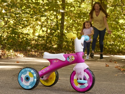 Young girl running towards the Huffy Mini Mouse electro-light trike with her mother smiling Young girl running towards the Huffy Mini Mouse electro-light trike with her mother smiling