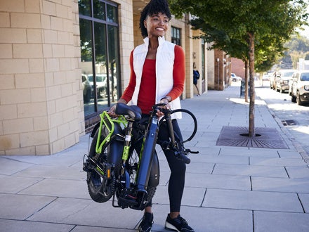 Woman carrying a folded Huffy Motoric electric bike on a city sidewalk, showcasing its portability and convenient design. Woman carrying a folded Huffy Motoric electric bike on a city sidewalk, showcasing its portability and convenient design.