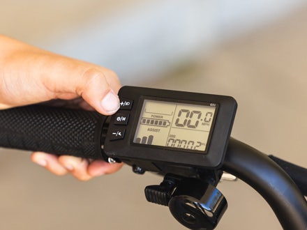 Close-up of a hand operating the Huffy Eventis e-bike display, showing speed, battery level, and power assist options on the console. Close-up of a hand operating the Huffy Eventis e-bike display, showing speed, battery level, and power assist options on the console.